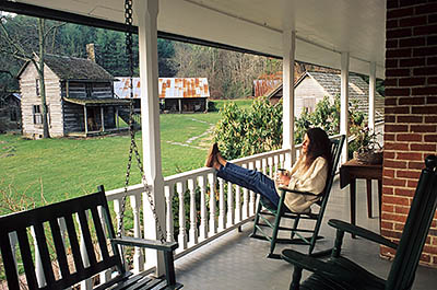 NC: Watauga County, Boone Area, Valle Crucis, The Mast Farm Inn, a Nat. Register site now a b&b. Deborah Bradshaw enjoying a morning cup of coffee on the front porch. Log outbldgs. in bkgd.  MOD. & PROP. REL. [Ask for #237.155.]